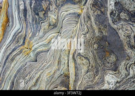 Close-up of metamorphic rock surface with flowing layered striations and mineral veins in gray, blue, and ochre tones, creating an abstract natural st Stock Photo