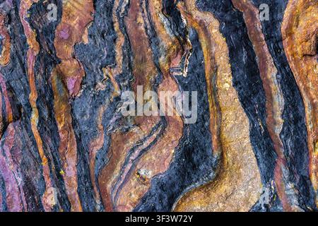Close-up of metamorphic rock texture with dramatic folded layers and mineral veins in blue, gold, and rust tones, forming an abstract stone background Stock Photo