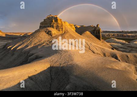 Wide-angle view of rugged sandstone badlands in the American Southwest, USA, lit by warm sunset light as a rain shower clears and a rainbow arcs over Stock Photo