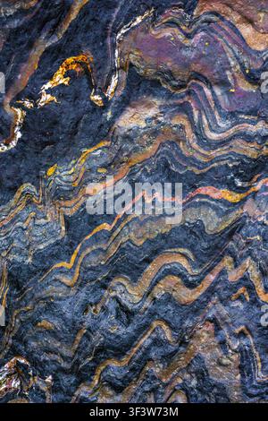 Abstract metamorphic rock surface texture with flowing wavy bands and mineral veins in dark blue, gray, and gold tones, ideal as a natural stone backg Stock Photo