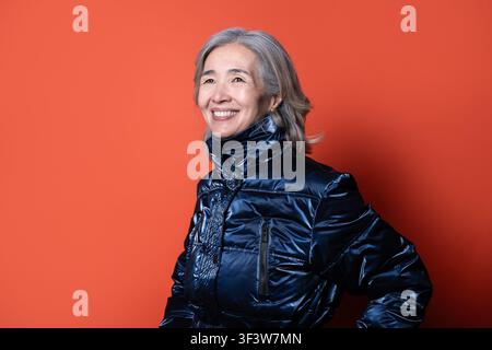 Studio portrait of a senior Asian woman smiling confidently, wearing a shiny navy puffer jacket, standing in profile against a bright coral background Stock Photo