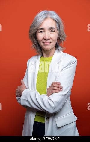 Confident senior Asian woman with gray hair wearing a white blazer and green top, arms crossed, posing in studio against a bold orange background. Stock Photo
