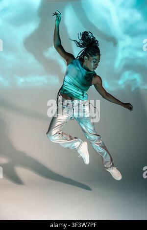 Afro woman dancer in dynamic midair jump, wearing silver pants and crop top, performing contemporary street dance in studio with blue projected light Stock Photo