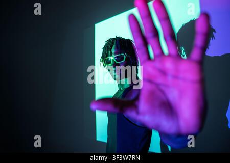 African man wearing sunglasses poses in neon cyan and purple lighting, raising his hand toward the camera for a bold futuristic portrait with deep sha Stock Photo