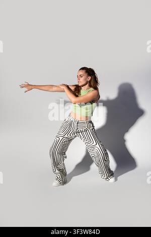 Urban woman dancer strikes an expressive pose in a green crop top and patterned pants against a gray studio wall, illuminated by strong light that cas Stock Photo