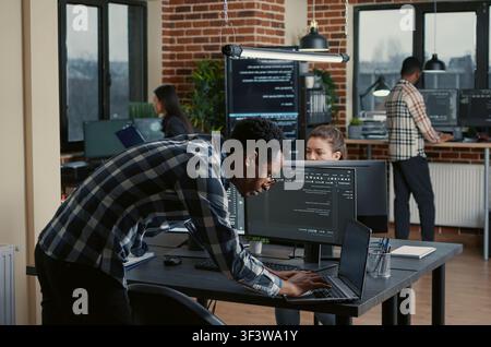 Senior developer codes on a laptop at a desk with multiple screens parsing software, compiling programming language and reviewing algorithm results. Stock Photo