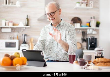 Authentic senior man on online video call in the morning in cozy kitchen, with elderly person using internet online chat technology video webcam making a video call connection camera communication. Stock Photo