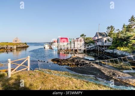 A fishing net stretched out like a fence frames the foreground of a view of Blue Rocks Harbour on The Point Road near Lunenburg, Nova Scotia, Canada. Boathouses with red tin roofs and a fishing boat are moored at low tide against the backdrop of the open Atlantic. Blue Rocks, Nova Scotia, Canada Stock Photo