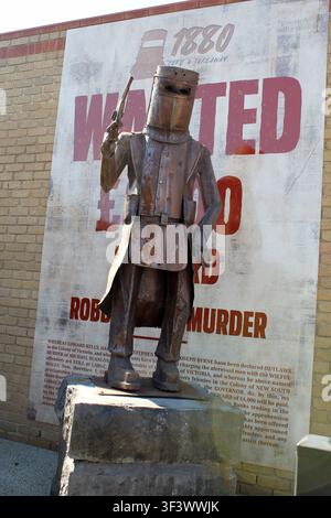 A weathered metal Ned Kelly statue holding a pistol, set against a vintage 1880 wanted poster billboard in Glenrowan, Victoria, Australia Stock Photo