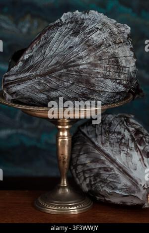 Two heads of dark purple cabbage with white bloom on the leaves, with an antique silver compote dish and a background of blue Venetian marbled paper. Stock Photo