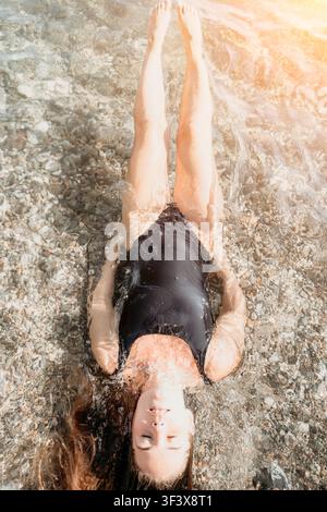 Woman water swimsuit young woman floats in clear shallow sea over pebbles with her legs in the air on a sunny summer day Stock Photo