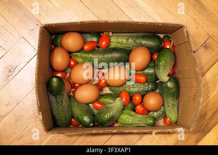 Cardboard box filled with a selection of raw, organic produce including fresh green cucumbers, small red cherry tomatoes, and brown eggs, sitting on a Stock Photo