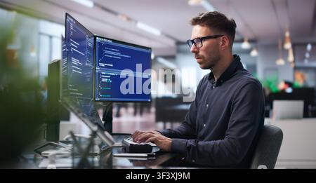 Young Software Engineer Works On Complex Algorithms Using Dual Screens At Desk. Stock Photo