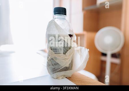 Plastic soda bottle wrapped in crumpled paper towel on wooden desk near window, soft natural light, Stock Photo