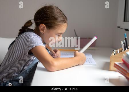 Happy elementary school girl sitting at a desk, reading her notebook while studying at home in a bright and modern workspace. Stock Photo