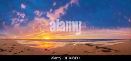 Sunset-lit clouds create a fiery panorama above tranquil water gently washing onto an empty beach Stock Photo