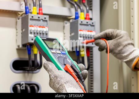 Electrician wearing protective gloves using digital multimeter to measure voltage in electrical control panel. Stock Photo