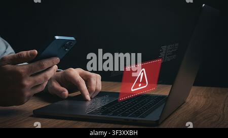 Cybersecurity breach concept with warning sign icon on laptop screen while user holding smartphone. Stock Photo