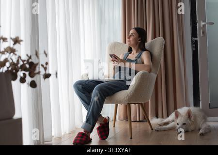 Young woman with a laptop and smartphone taking a break from working at home while a white pet dog rests beside her. Stock Photo