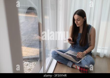 Young woman wearing a wireless earbud checks her smartphone while working on a laptop and sitting cross-legged beside a large window at home. Stock Photo