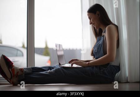 Smiling young woman wearing a wireless earbud works on a laptop while sitting on the floor with her legs extended beside a large window at home. Stock Photo