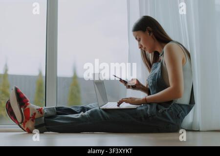 Focused young woman wearing a wireless earbud checks her smartphone while working on a laptop and sitting on the floor beside a large window at home. Stock Photo