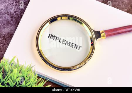 Magnifying glass rests on a clean white notepad displaying the word Implement clearly under the lens to emphasize focus on business strategies and pro Stock Photo