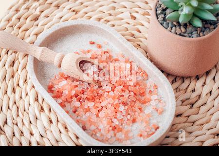 Pink Himalayan salt crystals in a ceramic dish with a wooden scoop and succulent on a woven placemat. Top view, flat lay. Stock Photo