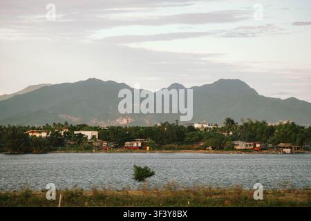 A wide river flows past a rural bank with houses and boats nestled among dense green trees, backed by massive dark mountain peaks under an overcast sk Stock Photo