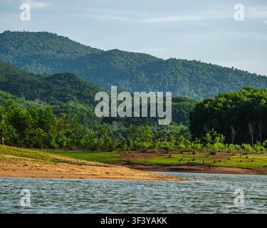 A wide view of a rippling body of water looking toward a sandy beach, green grass with small trees, and massive forested ridges under a soft pale sky. Stock Photo
