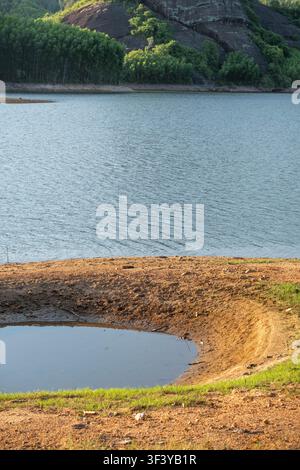 A small circular pool of water sits inside a deep explosion crater on a sandy and grassy bank next to a wide lake during a bright sunny day. Stock Photo