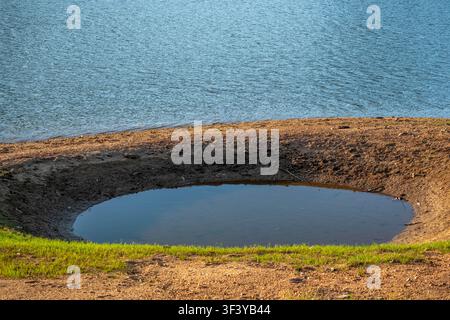 A small circular pool of water sits inside a deep explosion crater on a sandy and grassy bank next to a wide lake during a bright sunny day. Stock Photo
