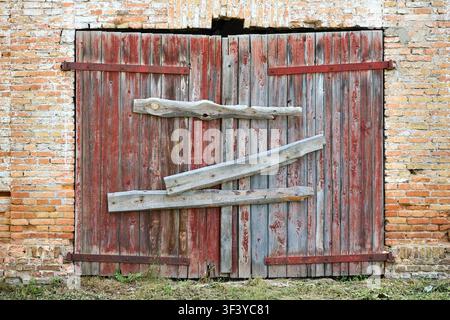 Weathered wooden barn door with peeling red paint boarded up with rough planks, set in old brick wall of abandoned rural building structure. Stock Photo