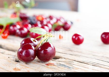 Two glossy cherries with stems and leaf rest on weathered wooden board, with blurred cherries behind, soft highlights, rich red color and fresh summer harvest feeling, with berry, summer, juicy Stock Photo