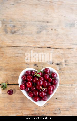 Close overhead view of glossy ripe red cherries overflowing heart shaped white bowl on weathered wooden table, with scattered leaves and one loose cherry, bright natural light creating fresh Stock Photo