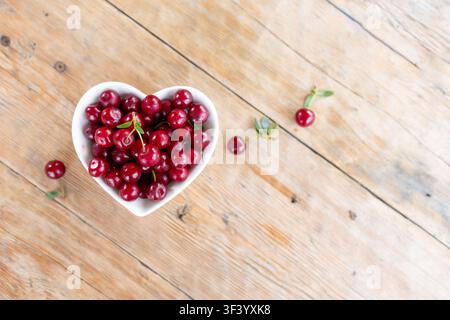 Top view of heart shaped white bowl overflowing with glossy ripe red cherries on weathered wooden plank table, few cherries and green stems scattered nearby, soft natural light, warm summer mood Stock Photo
