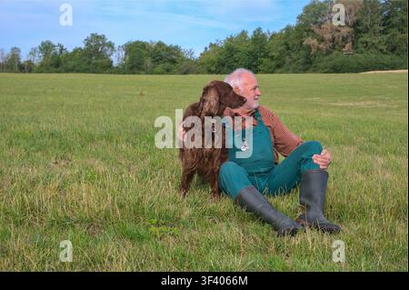 An elderly man wearing green dungarees and rubber boots sits in a grassy field next to his faithful Irish Setter dog, both looking into the distance. Stock Photo