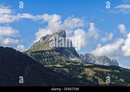 A massive Dolomite limestone peak rises above dark forested slopes under clear blue sky and scattered clouds in South Tyrol, northern Italy. Telephoto Stock Photo