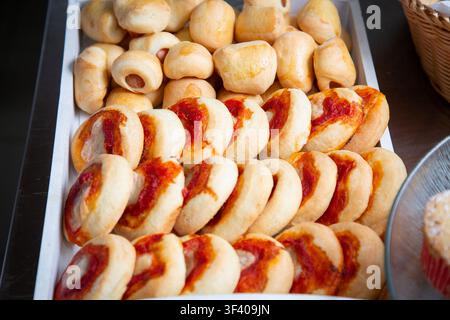Tray of assorted baked bread rolls with golden crust and red sauce topping, arranged in close packed rows for bakery display, buffet service, snack Stock Photo