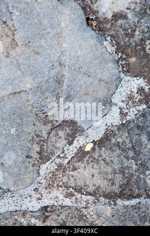 Weathered gray stone surface with cracks, rough mineral texture, pale vein lines, dark patches, and worn edges forming abstract natural background for Stock Photo