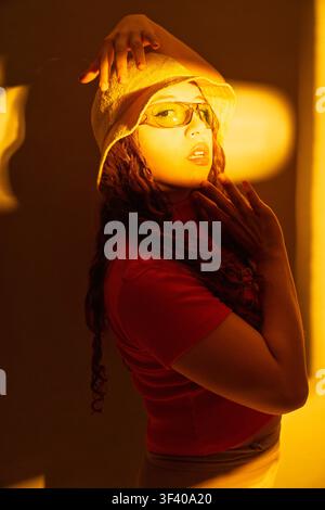 Young woman wearing a bucket hat and sunglasses posing with hands, illuminated by warm orange light Stock Photo
