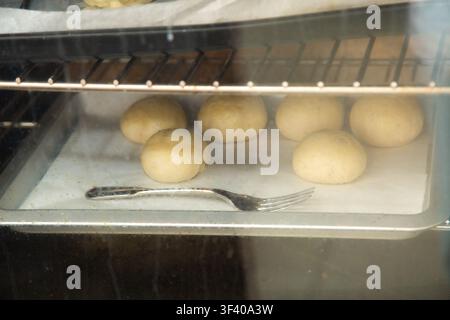 Bread dough balls rising on parchment inside oven with metal fork on tray, warm bakery scene showing proofing process, soft light, rustic home baking Stock Photo