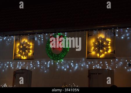 a beautifully illuminated Christmas wreath adorns the wooden shutters of a house exterior at night, featuring string lights and festive green and red Stock Photo