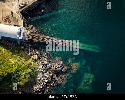 Aerial top down view directly above an old British lifeboat ramp on the Cornish coast sloping down into the sea with a small rowing boat in a Cornwall Stock Photo