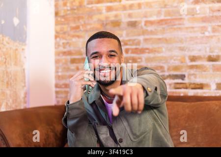 Smiling young man pointing finger while talking on phone Stock Photo