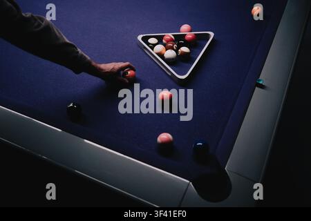 Player’s hand positions a billiard ball beside a triangle rack on a dark blue pool table in a dimly lit game room Stock Photo
