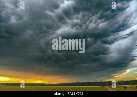 Dramatic mammatus storm clouds forming above a rural landscape during sunset. Heavy rainfall in the distance, colorful evening sky, severe weather Stock Photo