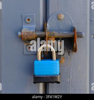 Blue padlock secures weathered metal door latch. Rust and wear mark the aged lock mechanism. The padlock's vibrant color contrasts the grey wood. It s Stock Photo