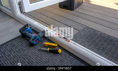 Tools rest near a sliding glass door frame. Tools lie on a dark mat beside a wooden deck. Bright sunlight glows through the glass. Work appears paused Stock Photo