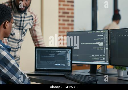 Software developer writes code on a laptop while a coworker asks for advice, showing programming teamwork and cloud computing collaboration. Stock Photo